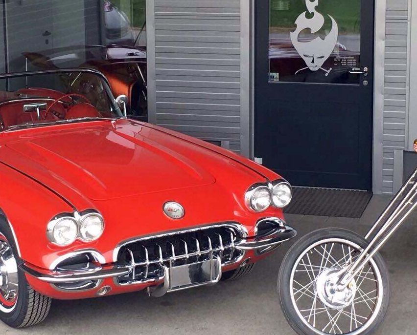 Ein roter Vintage Chevrolet Corvette Cabriolet ist neben einem Motorrad vor einem grauen Gebäude mit einer Glastür geparkt.