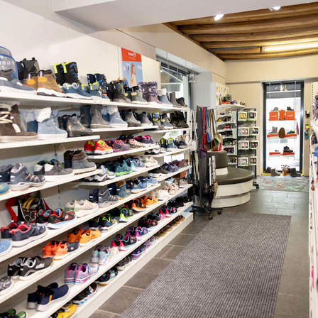 Ein Schuhgeschäft mit Regalen, die mit verschiedenen Schuhen, einschließlich Stiefeln und Sneakern, gefüllt sind. Die Regale sind mit verschiedenen Farben und Stilen gefüllt. Es gibt einen Teppich auf dem Boden und eine Glastür, die zum Eingang führt.