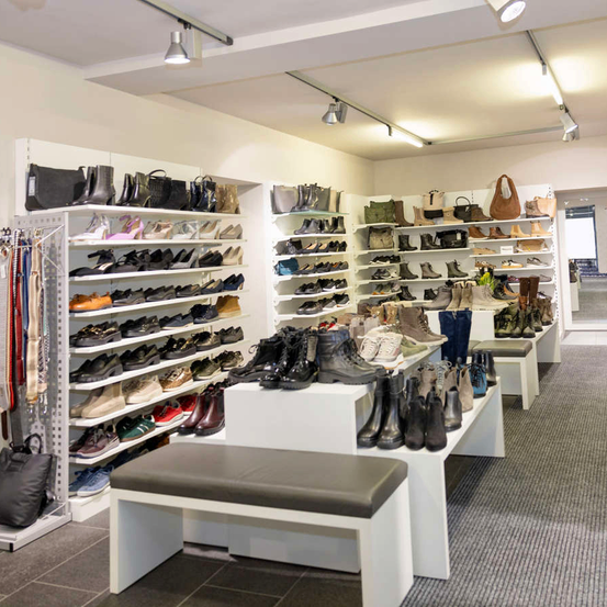 A shoe store displays a variety of shoes on shelves and benches. The room is well-lit with ceiling lights and has a clean and organized appearance.