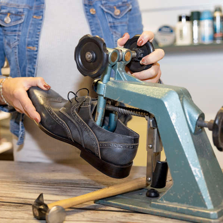 Eine Frau in einer Jeansjacke benutzt einen Schuhstrecker, um einen schwarzen Lederschuh auf einem Holztisch anzupassen, mit einem Hammer in der Nähe.