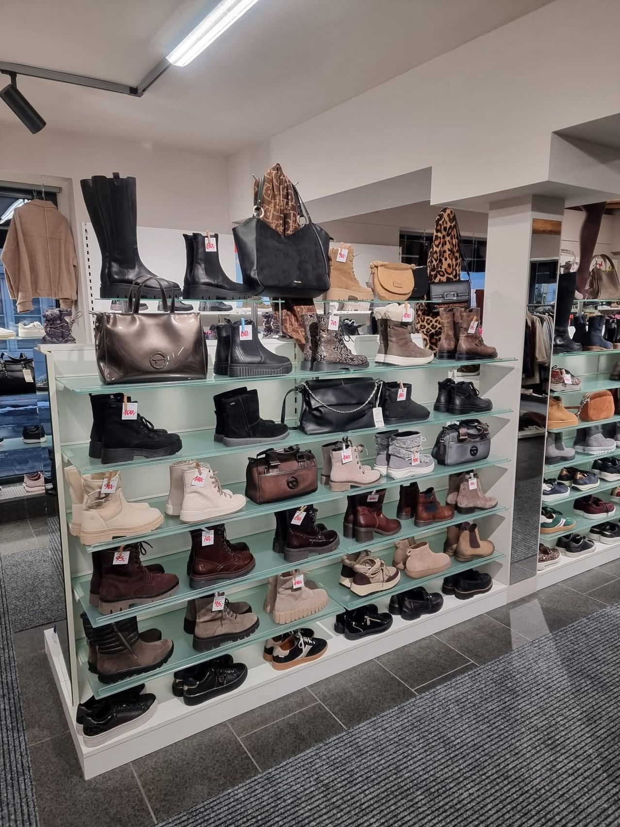 Ein Schuhgeschäft zeigt verschiedene Stiefel und Schuhe auf Glasregalen. Verschiedene Handtaschen sind ebenfalls ausgestellt. Preisschilder sind an den Artikeln befestigt.