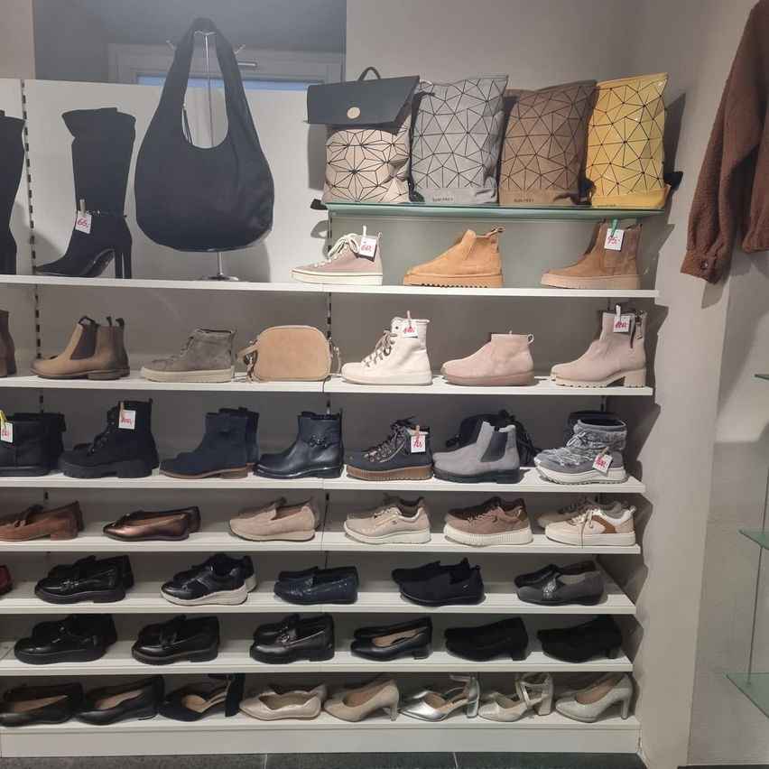 Ein weißer Regalboden zeigt verschiedene Schuhe, darunter Stiefel, Sneaker und elegante Schuhe. Über dem Regal hängen Taschen an einem Ständer. Die Schuhe sind in verschiedenen Farben und Stilen.