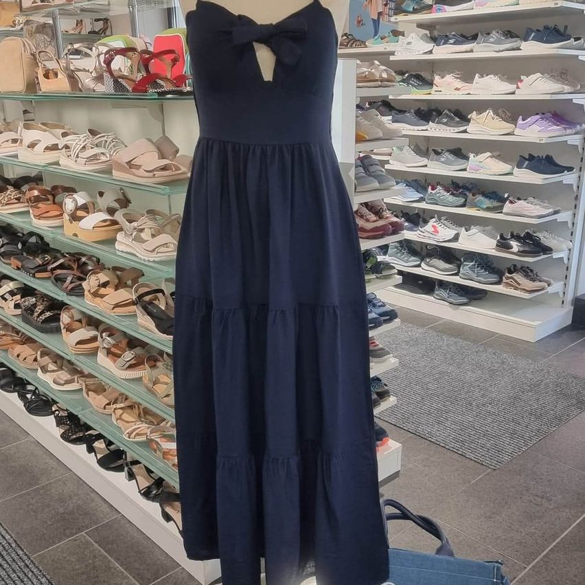 Ein dunkelblaues Kleid mit einer Schleife am Ausschnitt wird in einem Schuhgeschäft neben vielen Paar Schuhen auf Regalen ausgestellt.