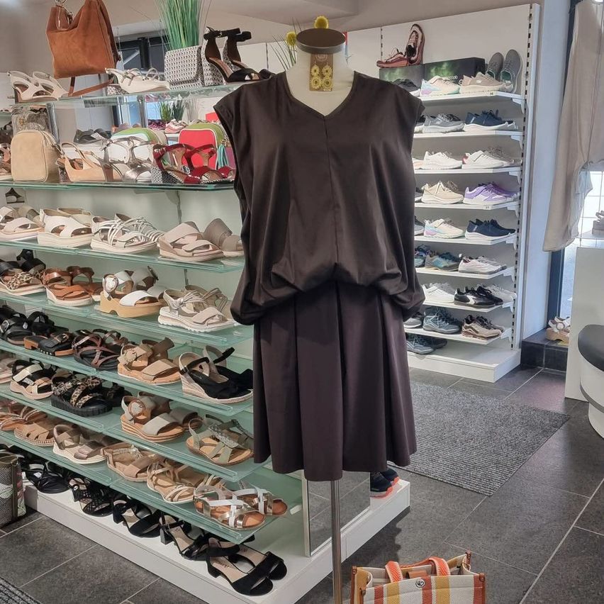 Ein braunes ärmelloses Kleid wird auf einem Ständer in einem Geschäft ausgestellt, das mit verschiedenen Sandalen in den Regalen gefüllt ist.