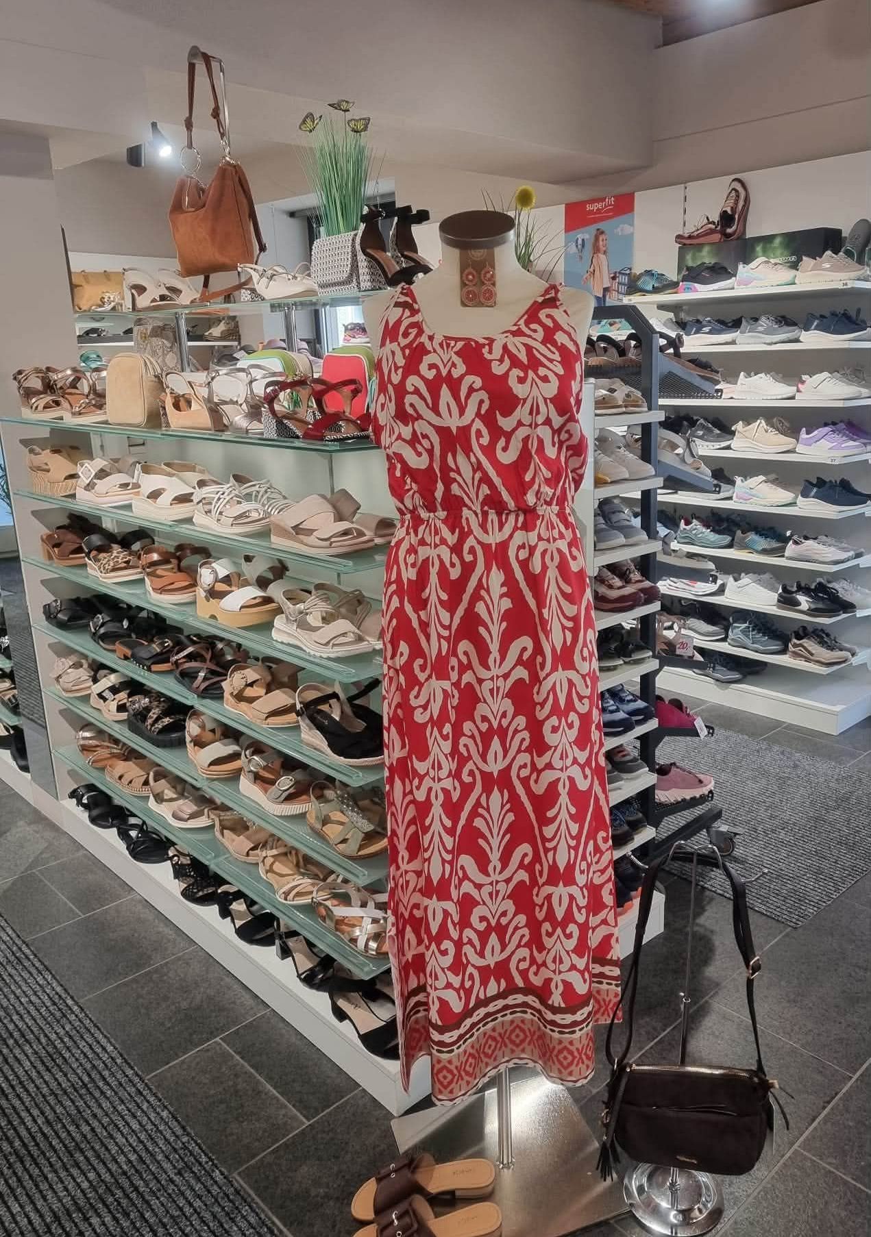Ein Schuhgeschäft mit einem roten Kleid auf einem Mannequin zwischen verschiedenen Schuhen. Regale zeigen Sandalen und Schuhe, mit einem Teppich auf dem Boden.