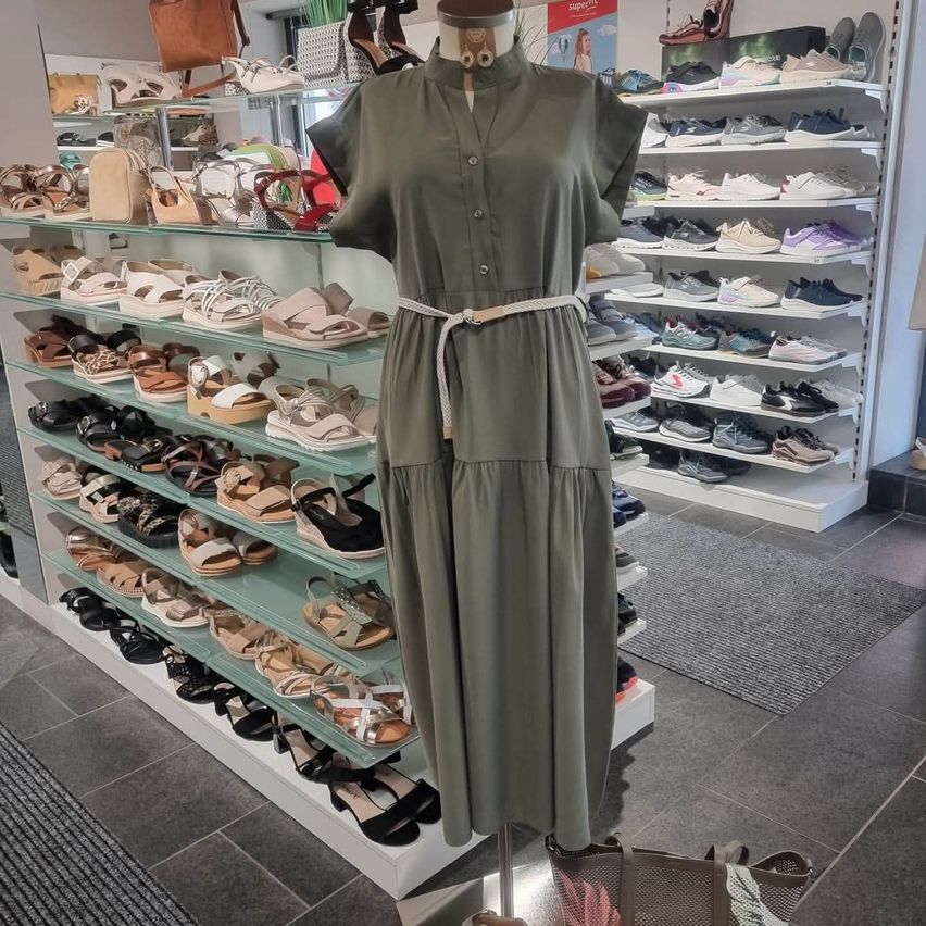 Ein Dummy präsentiert ein grünes Kleid in einem Geschäft, das mit verschiedenen Sandalen auf Regalen gefüllt ist.