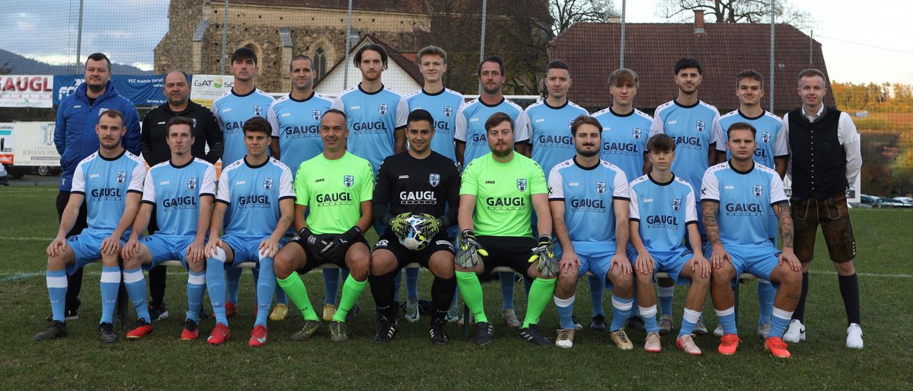 Eine Fußballmannschaft posiert für ein Gruppenfoto vor einer Kirche mit Turm und Zaun. Sie tragen blaue und weiße Uniformen mit Logo.