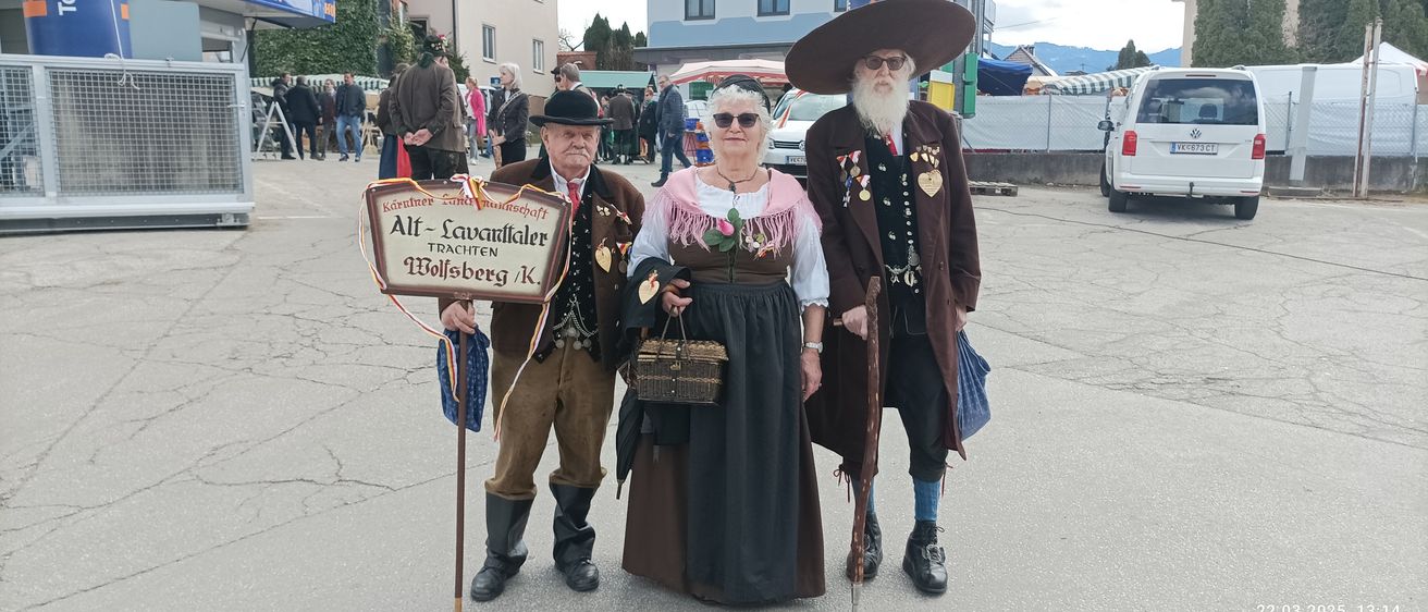 Drei Personen in traditioneller Kleidung stehen in einer Reihe. Ein Mann hält ein Schild, eine Frau einen Korb und ein weiterer Mann einen Stock. Sie tragen Hüte, Sonnenbrillen und Stiefel. Im Hintergrund sind Gebäude und Menschen zu sehen.