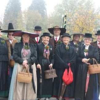 Eine Gruppe von Menschen in traditioneller Kleidung steht draußen und hält verschiedene Gegenstände wie Handtaschen, Regenschirme und Körbe. Sie befinden sich in einem nebligen Bereich mit Bäumen und Autos im Hintergrund.