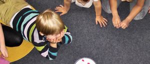 Drei kleine Kinder spielen auf einem Teppichboden. Ein Kind bedeckt sein Gesicht mit den Händen. Ein anderes Kind streckt die Hände aus. Ein drittes Kind ist hinter ihnen.