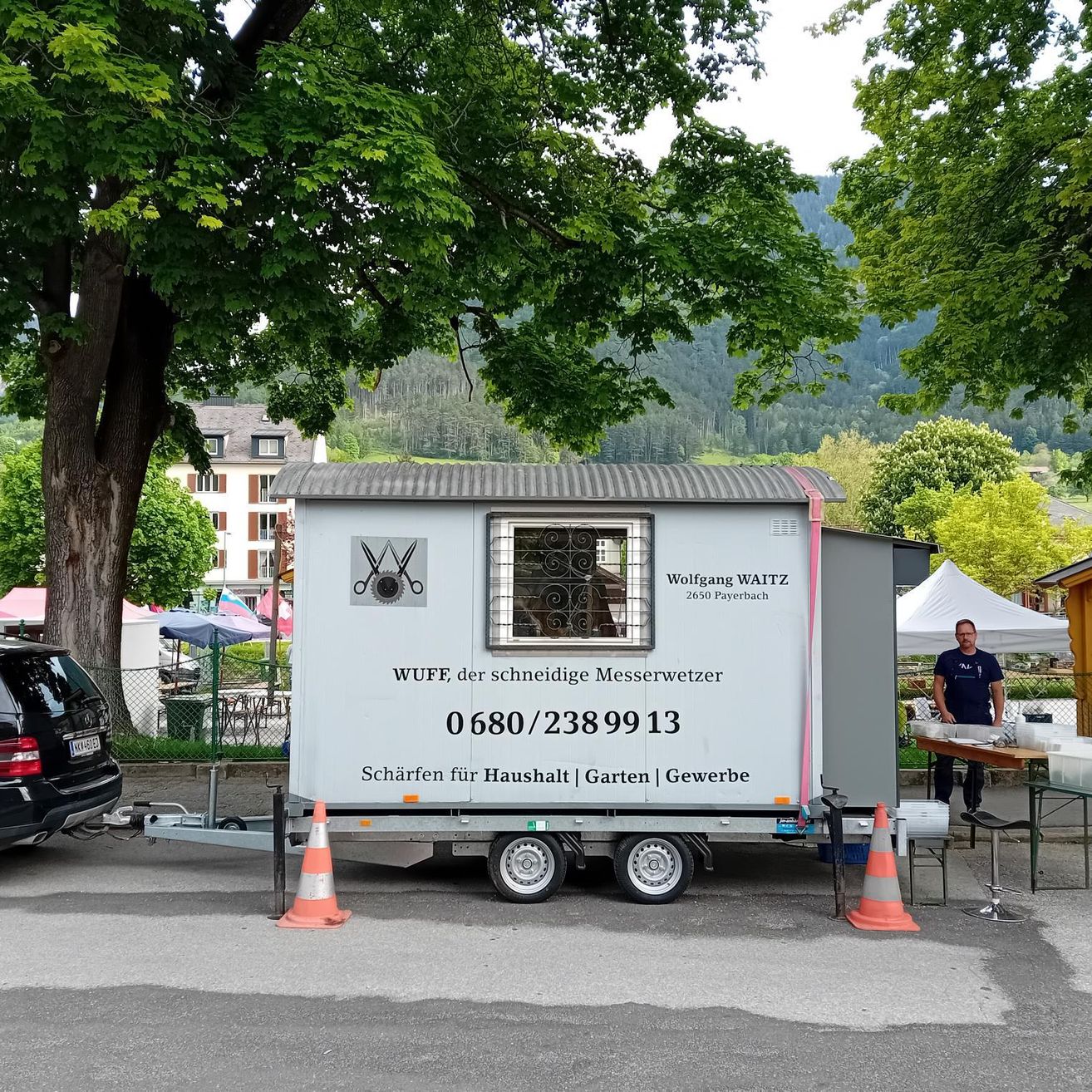 Ein weißer Anhänger mit dem Namen Wolfgang Waitz steht auf der Straße. Ein Mann steht neben einem Tisch mit Lebensmittelbehältern. Ein Auto und ein Baum sind in der Nähe.