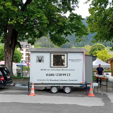 Ein weißer Anhänger mit dem Namen Wolfgang Waitz steht auf der Straße. Ein Mann steht neben einem Tisch mit Lebensmittelbehältern. Ein Auto und ein Baum sind in der Nähe.