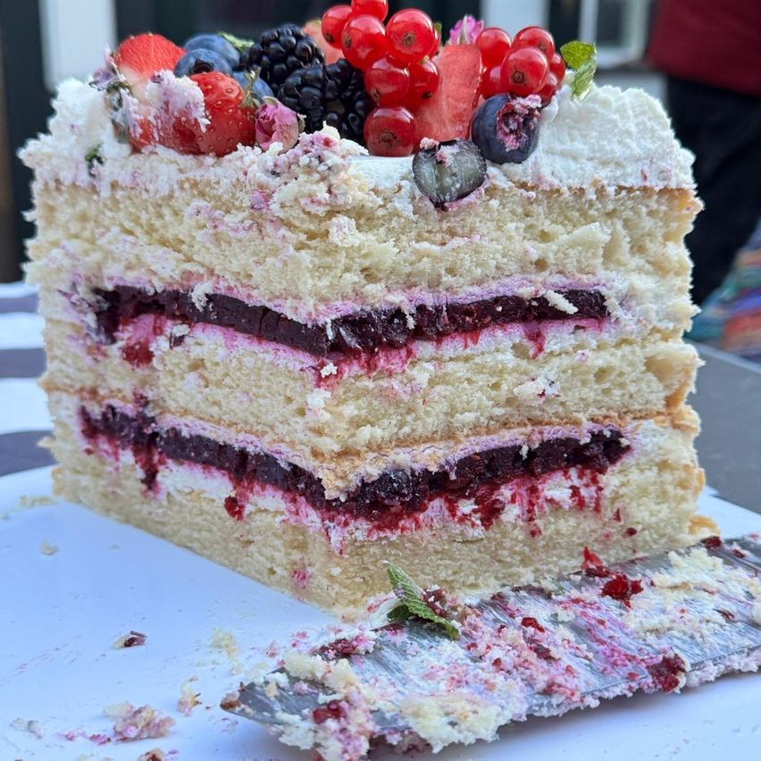 Ein Kuchenstück mit mehreren Schichten, gefüllt mit Beerenmarmelade, überzogen mit weißer Glasur und frischen Beeren, auf einem weißen Teller mit einem Messer abgelegt.