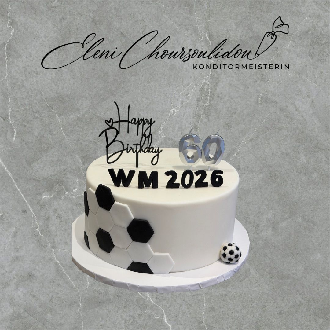 Ein weißer Kuchen mit einem Fußballdesign, der 'Happy Birthday WM 2026' darauf steht. Der Kuchen ist auf einem weißen Teller auf einer grauen Oberfläche.
