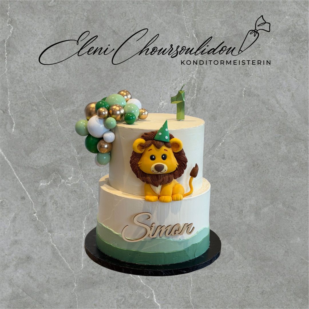 Ein zweistöckiger Kuchen mit einer Löwenfigur und Luftballons auf der Spitze, mit der Nummer 1 und dem Namen 'Simon' darauf geschrieben.
