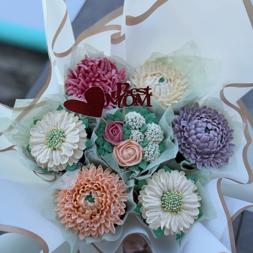 A bouquet of edible flowers in various colors, including pink, white, and purple, arranged in a white paper wrapper with a red heart-shaped tag that says 'Best Mom'.