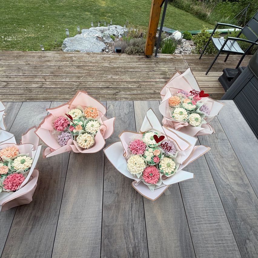 Three cupcake bouquets with pink and white flowers, hearts, and pink ribbons on a wooden deck. Behind it is a green lawn with plants.