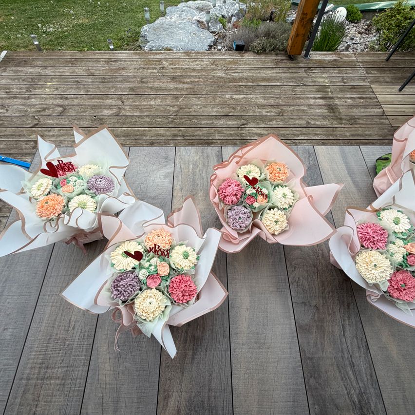 Three bouquets of cake flowers are arranged on a wooden deck, with pink wrappings and a mix of colors. The background shows a grassy area with rocks and plants.