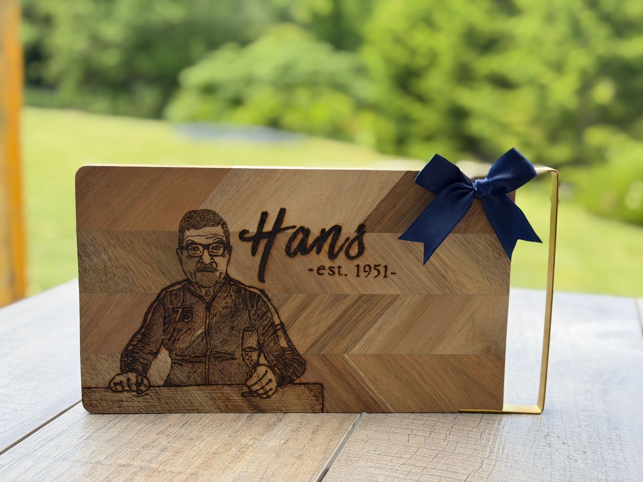 A wooden plaque with a drawing of a man wearing glasses and a jacket with the number 75. The plaque is tied with a blue ribbon and has the name Hans and the years 1951- written on it.