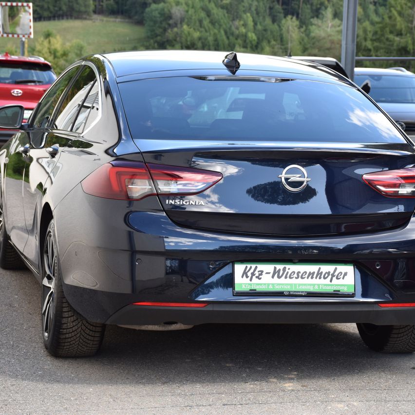 Ein schwarzer Opel Insignia steht auf einem Parkplatz mit einem Kennzeichen 'Kfz-Wiesenhofer.' In der Nähe sind weitere Autos geparkt.
