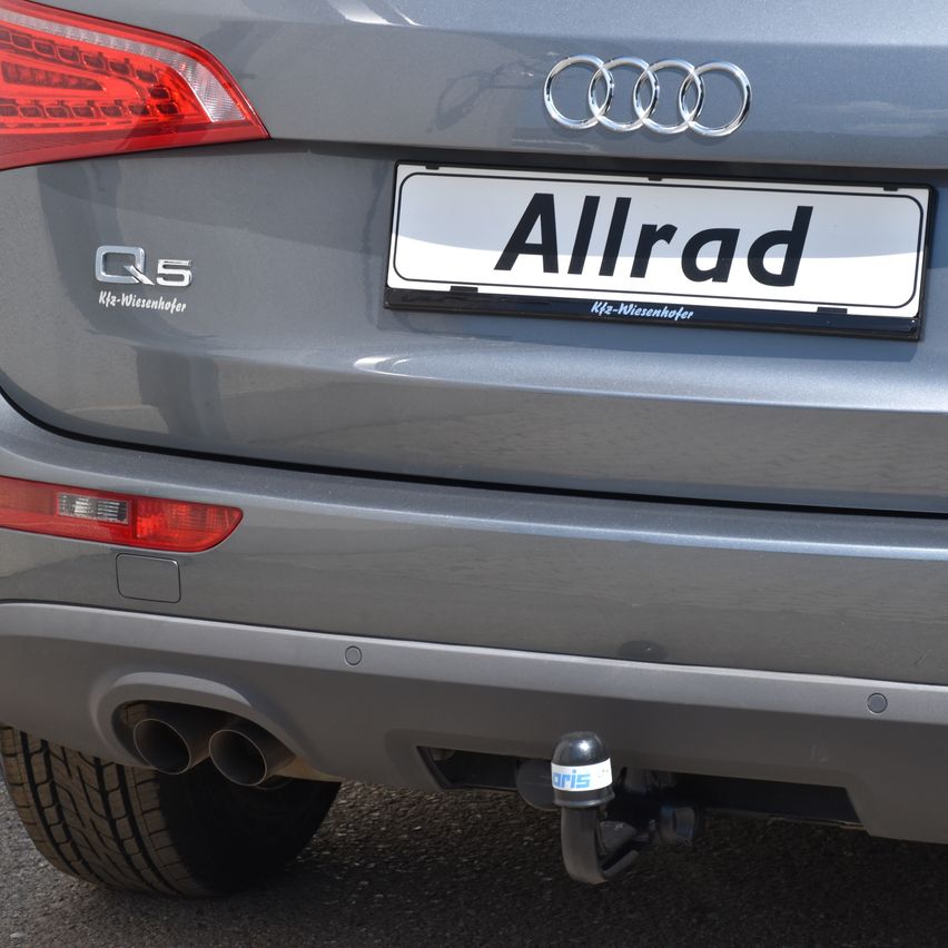Ein grauer Audi Q5 mit einem weißen Nummernschild, auf dem 'Allrad' steht, ist auf der Straße geparkt. Das Auto hat zwei Auspuffrohre und eine Anhängerkupplung. Das Audi-Logo befindet sich über dem Nummernschild.