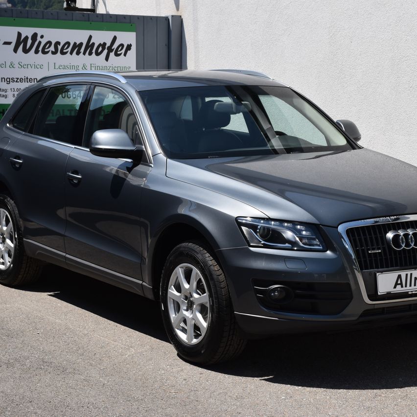 Ein grauer Audi SUV ist vor einer weißen Wand geparkt und zeigt sein schlankes Design. Das Auto trägt das Kennzeichen 'Allr'. Dahinter steht ein Schild mit dem Wort '-Wiesenhofer' in fetten Buchstaben.