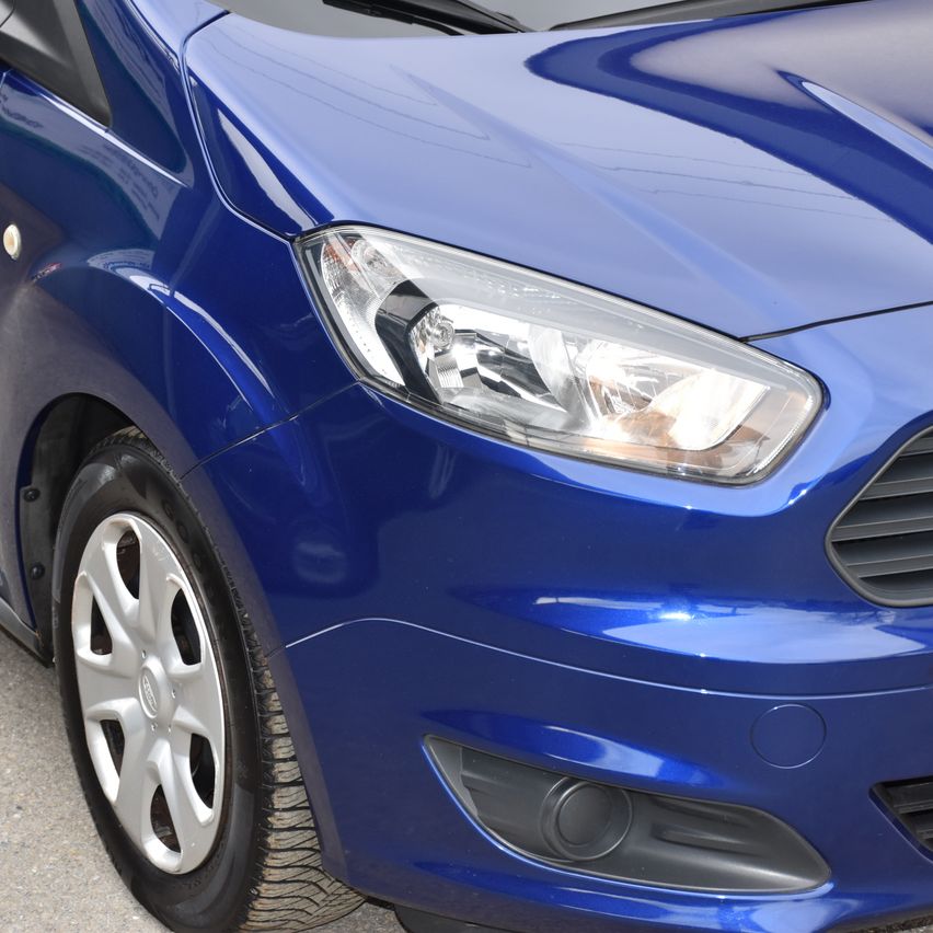 Eine Nahaufnahme der Vorderseite eines blauen Autos, mit Fokus auf Scheinwerfer und Stoßfänger. Der Scheinwerfer ist eingeschaltet und beleuchtet die Umgebung.