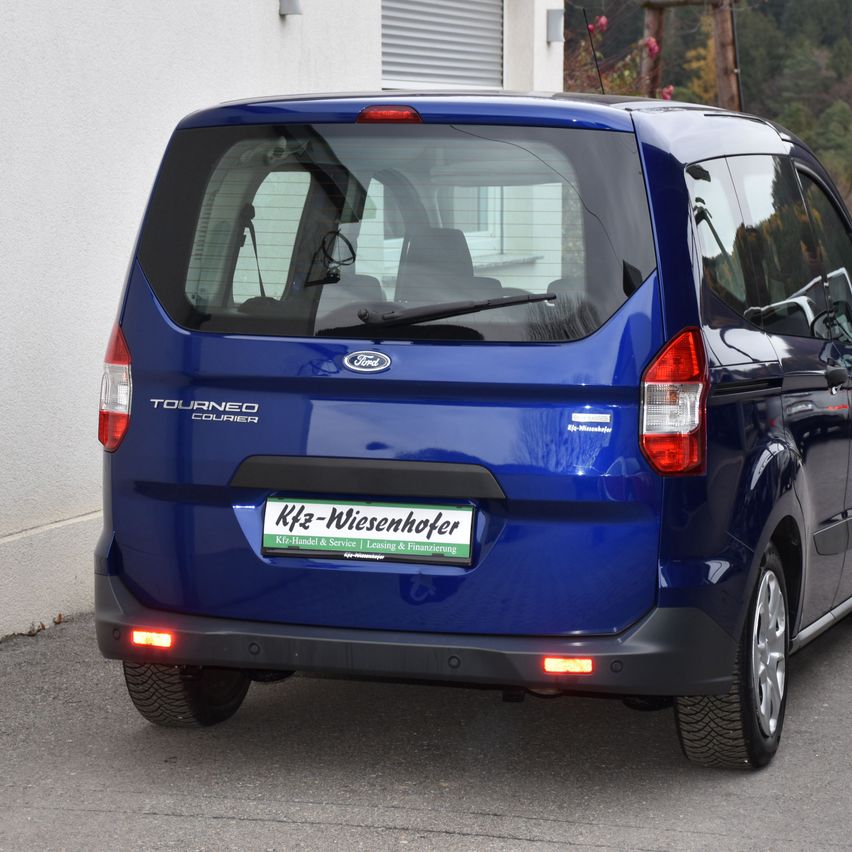 Die Heckseite eines blauen Ford Tourneo Courier steht am Straßenrand, mit roten Rücklichtern und einem Nummernschild, das 'Kfz-Wiesenhofer' liest.