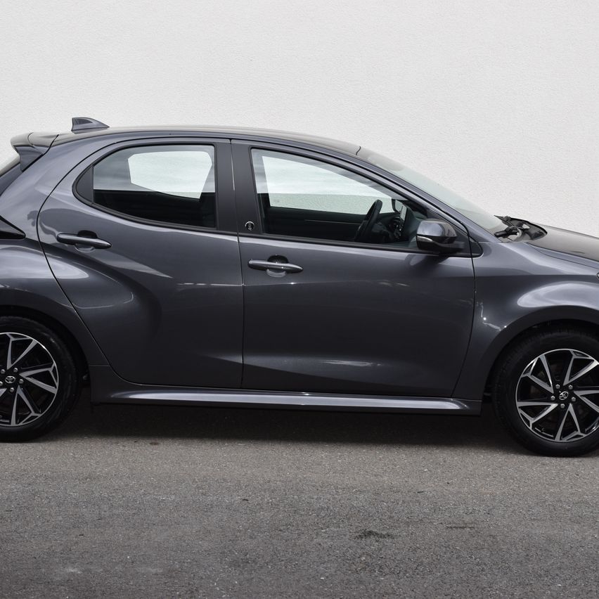 Ein grauer Toyota-Hatchback ist an einer weißen Wand geparkt. Das Auto hat ein schlankes Design mit einem gebogenen Spoiler und modernen Leichtmetallrädern.