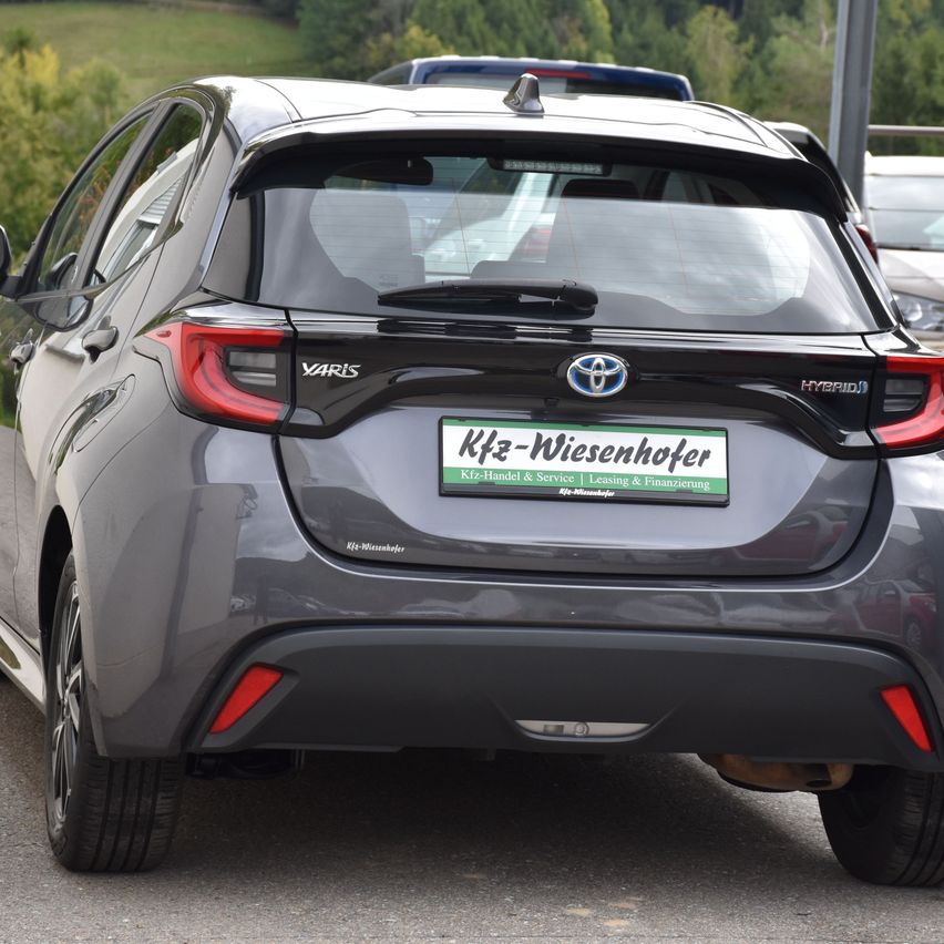 Die Heckseite eines grauen Toyota Yaris Hybridautos steht auf einer Straße mit einem Nummernschild, das 'Kfz-Wiesenhofer' liest.