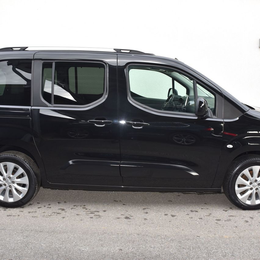 Ein schwarzer Minivan ist vor einer weißen Wand geparkt. Der Van hat vier Türen und drei Fenster. Die Reifen sind schwarz.