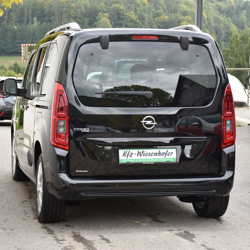 Die Heckansicht eines schwarzen Opel Combo-Vans ist auf einer Straße mit einem grünen Nummernschild geparkt. Bäume und ein Gebäude sind im Hintergrund zu sehen.
