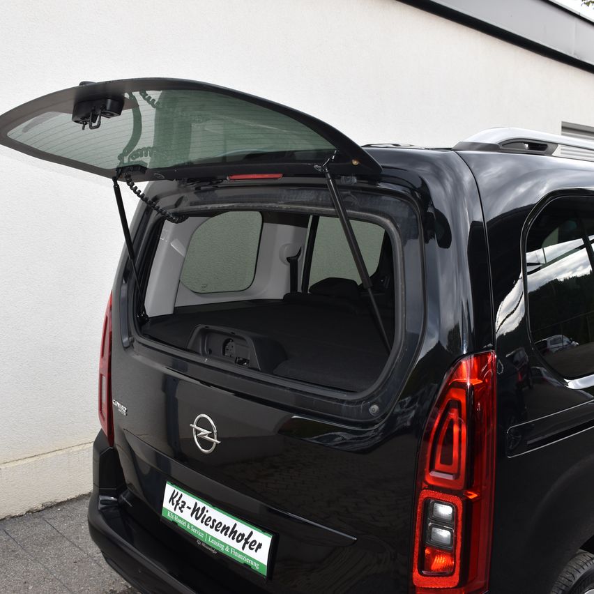 Die Rückseite eines schwarzen Opel-Vans mit offenem Kofferraum, der Innenraum und ein Nummernschild zeigt. Der Van ist gegen eine weiße Wand geparkt.