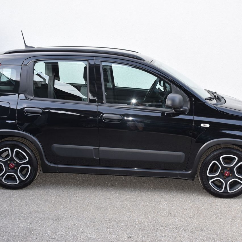 Ein schwarzer Fiat 500L mit schwarzen Reifen und Leichtmetallrädern steht auf einem Betonuntergrund vor einer weißen Wand.