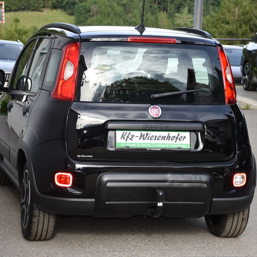 Die Rückseite eines schwarzen Fiat Panda, der auf einer Straße geparkt ist. Das Auto hat ein Nummernschild mit grüner Schrift, rote Rückleuchten und einen Ersatzreifen hinten.