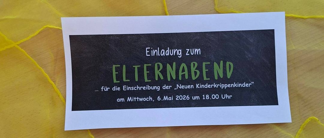 Eine weiße Karte mit schwarzem Hintergrund. Es steht: 'Einladung zum ELTERNABEND...für die Einschreibung der 'Neuen Kinderkrippenkinder' am Mittwoch, 6. Mai 2026 um 18.00 Uhr'. Die Karte liegt auf einem gelben Tuch.