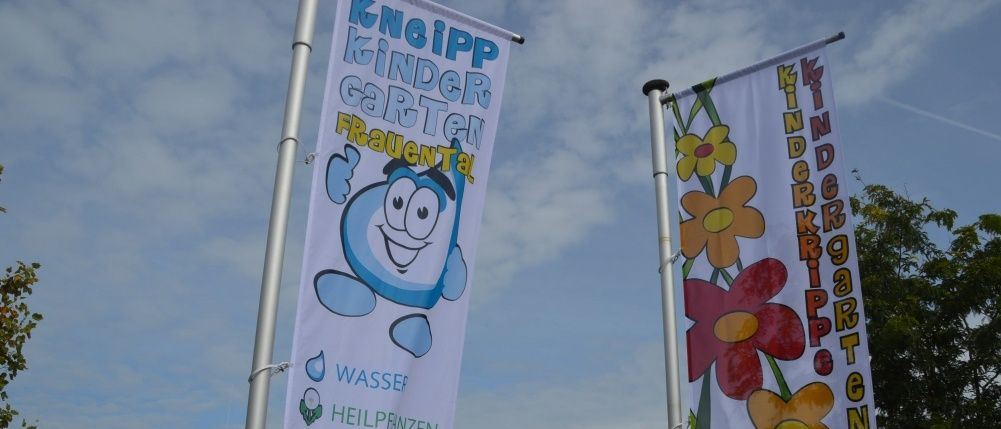 Zwei Banner mit Comicfiguren und Blumen sind vor einem Gebäude mit Solarmodulen auf dem Dach ausgestellt. Ein Banner zeigt 'Kneipp Kindergarten Frauental' und listet Dienstleistungen wie Wasser, Pflege, Ernährung, Bewegung und Lernfreude auf.