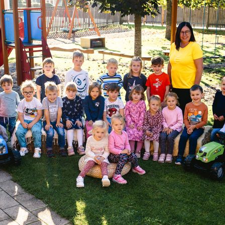 Eine Gruppe von Kindern und einer Frau steht in einem Park. Die Kinder sitzen auf Heuballen und Spielzeug. Hinter ihnen sind ein Rutsch und andere Spielgeräte. Die Frau steht vor ihnen.
