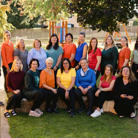 Eine Gruppe von Frauen in verschiedenen Farben von Hemden und Hosen posiert für ein Foto in einem Park mit Bäumen und einem Spielplatz im Hintergrund.