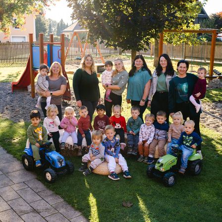 Eine Gruppe von Frauen und kleinen Kindern posiert für ein Foto in einem Spielplatz mit Spielzeugtraktoren und einer Rutsche im Hintergrund.