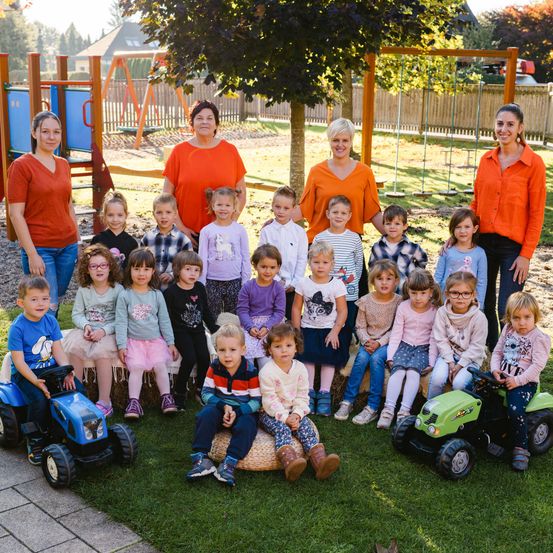 Eine Gruppe von Kindern und drei erwachsenen Frauen stehen mit Spielzeug vor einem Spielplatz auf einem Rasen.