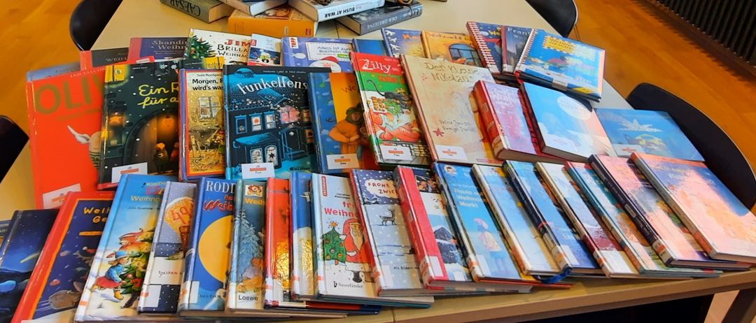 Ein langer Tisch in einem Raum mit Büchern in einer Pyramidenform angeordnet, dekoriert mit Tannenzapfen und Grün. Neben dem Tisch stehen Stühle und ein Monitor. Draußen am Fenster ist ein Auto zu sehen.