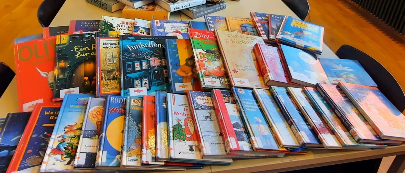 Ein langer Tisch in einem Raum mit Büchern in einer Pyramidenform angeordnet, dekoriert mit Tannenzapfen und Grün. Neben dem Tisch stehen Stühle und ein Monitor. Draußen am Fenster ist ein Auto zu sehen.