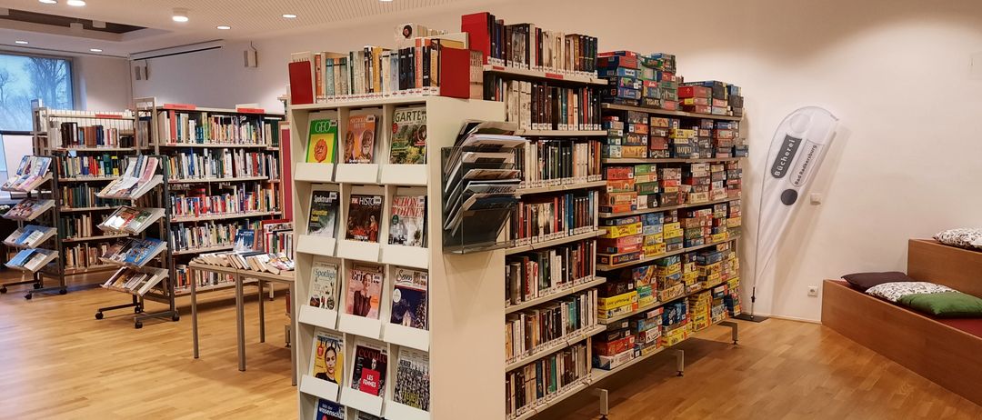 Eine Bibliothek mit mehreren Regalen voller Bücher und Zeitschriften. Ein Tisch hält Bücher. Ein anderes Regal hält Brettspiele.
