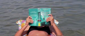 Eine Frau in einem schwarzen Bikini schwimmt auf einem bunten aufblasbaren Schlauchboot im Wasser und liest ein Buch, wobei ihr Gesicht dahinter verborgen ist.
