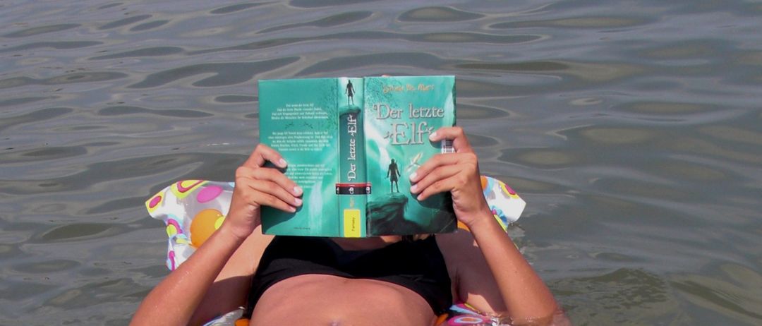 Eine Frau in einem schwarzen Bikini schwimmt auf einem bunten aufblasbaren Schlauchboot im Wasser und liest ein Buch, wobei ihr Gesicht dahinter verborgen ist.