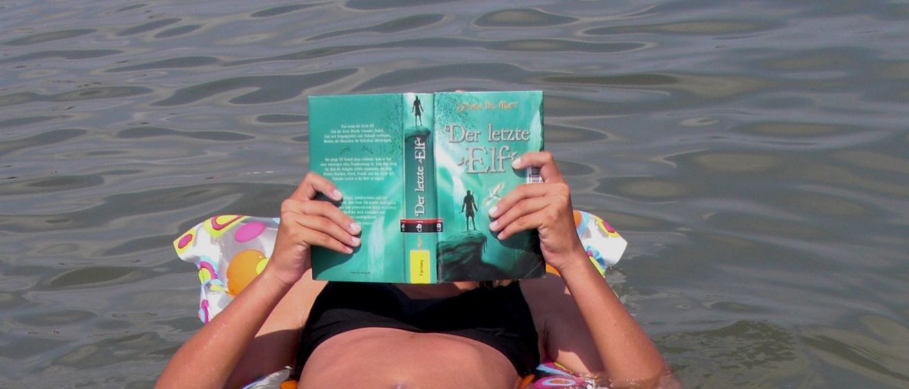 Eine Frau in einem schwarzen Bikini schwimmt auf einem bunten aufblasbaren Schlauchboot im Wasser und liest ein Buch, wobei ihr Gesicht dahinter verborgen ist.