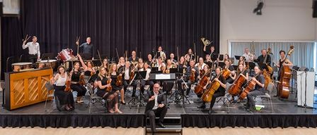 Ein Symphonieorchester auf der Bühne mit Musikern, die verschiedene Instrumente spielen, einschließlich Cellos, Violinen und Trommeln, unter Bühnenlicht in einem Theater.
