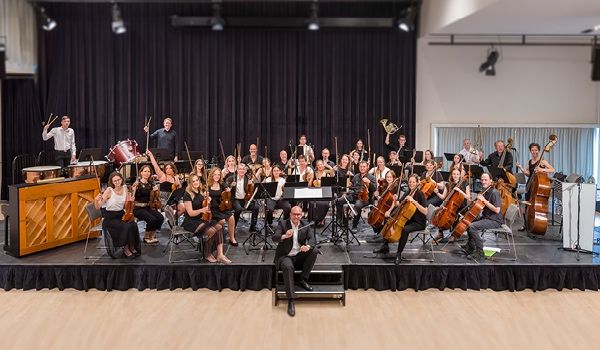 Ein Symphonieorchester auf der Bühne mit Musikern, die verschiedene Instrumente spielen, einschließlich Cellos, Violinen und Trommeln, unter Bühnenlicht in einem Theater.