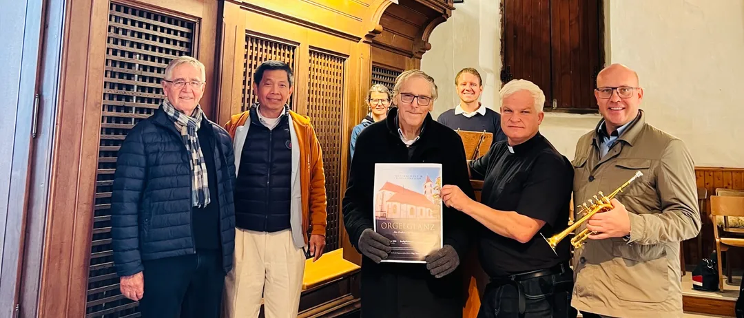 Fünf Männer stehen vor einer Kirchenorgel. Ein Mann hält ein Buch und ein anderer hält eine Trompete.